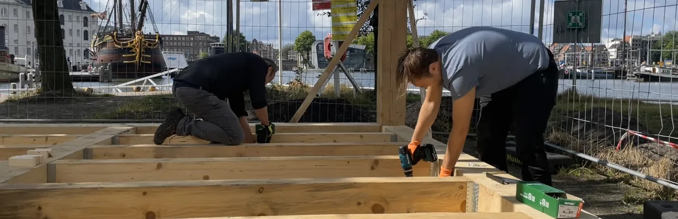Two people kneeling on the timber deck of the pavilion, assembling the structure by hand