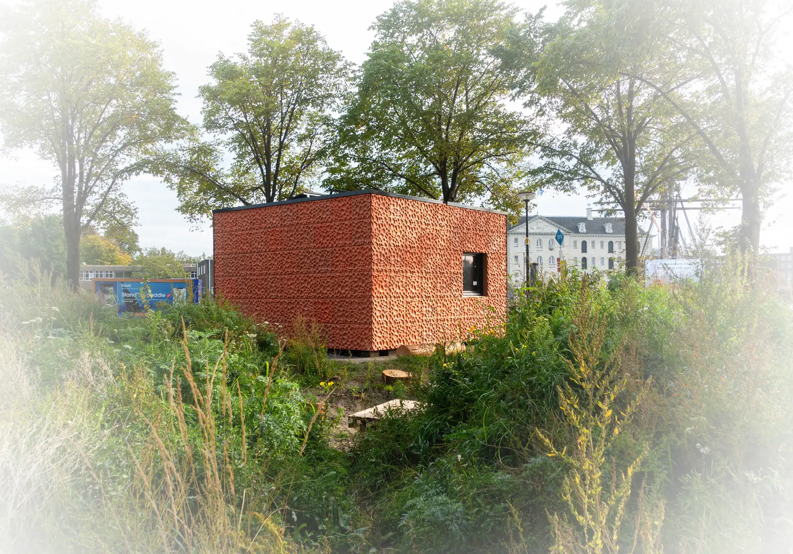 The completed pavilion standing among tall grasses and trees, clad in textured red facade panels